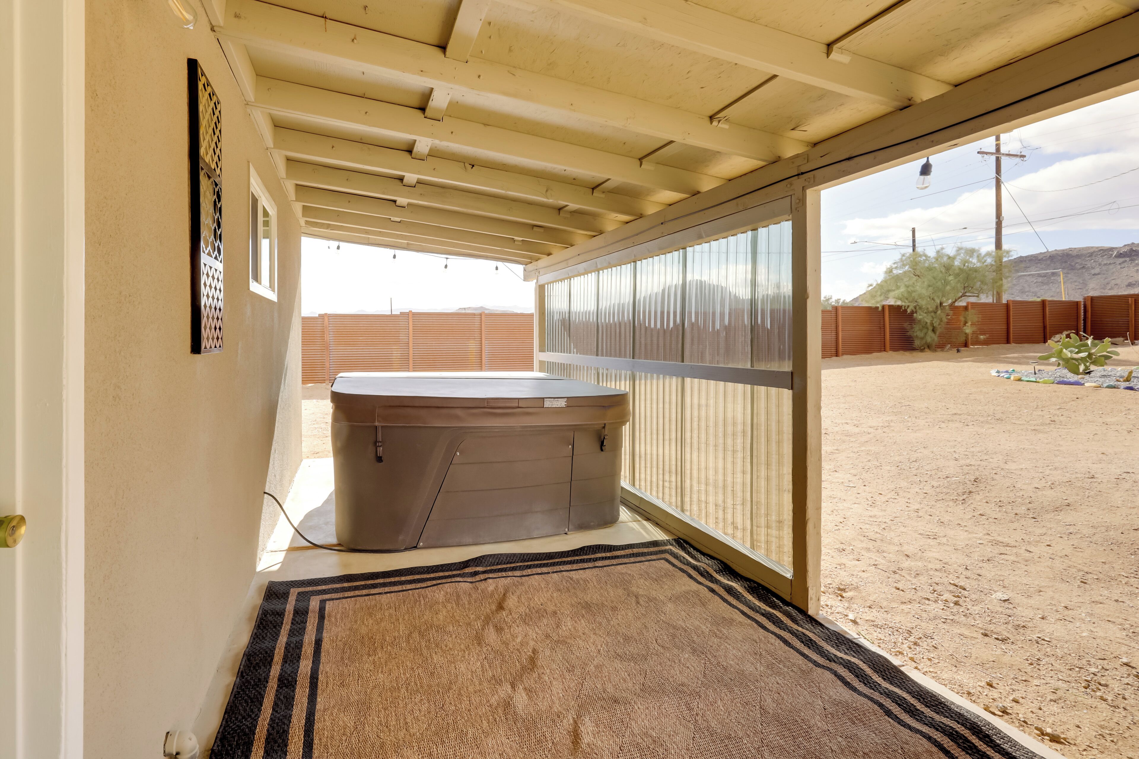 A hot tub under a covered patio with a view of a fenced yard and desert landscape.