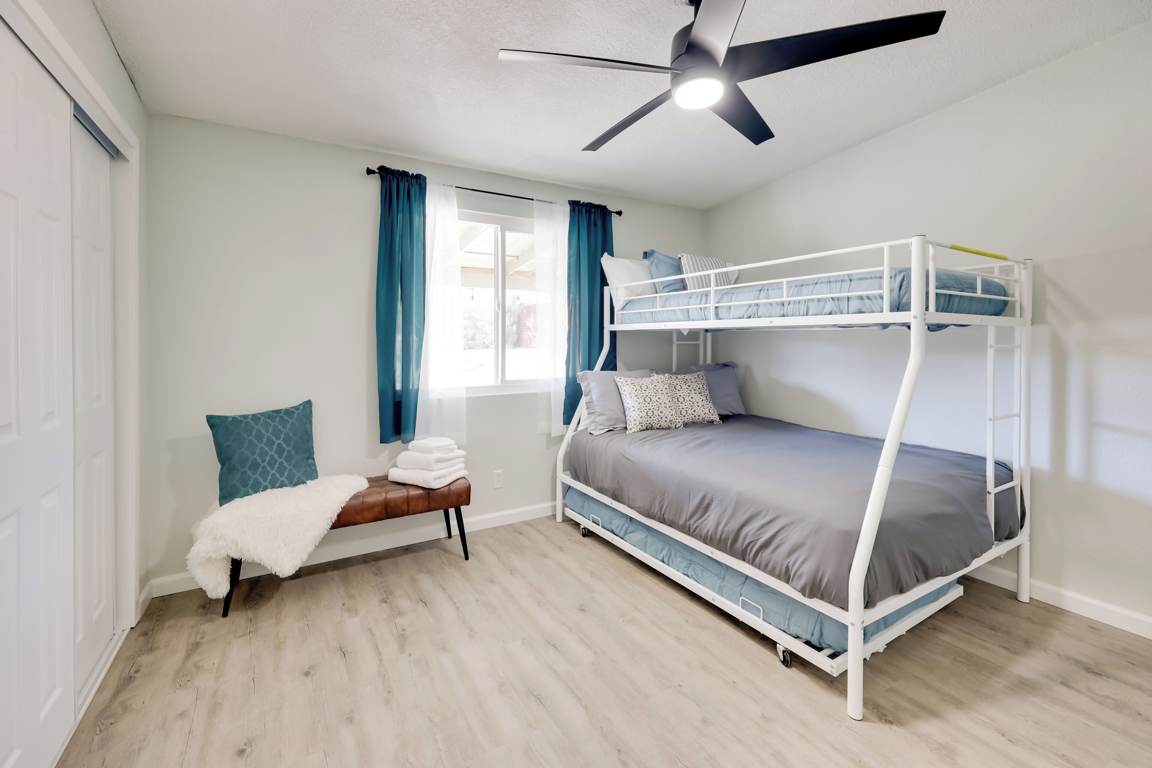 Une chambre avec un lit superposé, un ventilateur de plafond, une fenêtre avec des rideaux et un banc avec une couverture.