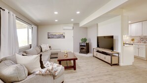 A modern living room with a sofa, coffee table, TV, and a kitchen area with cabinets. - Mtn-view Home w/ Hot Tub: 8 Mi to Joshua Tree! (Joshua Tree)