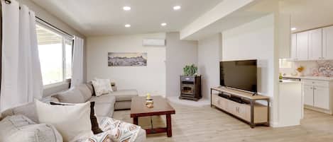 Una sala de estar moderna con sofá, mesa de centro, televisor y una zona de cocina con armarios.