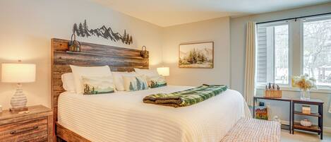 Un dormitorio con cabecera de madera, una cama grande con ropa blanca, una mesita de noche con lámpara y una ventana con vista a árboles.