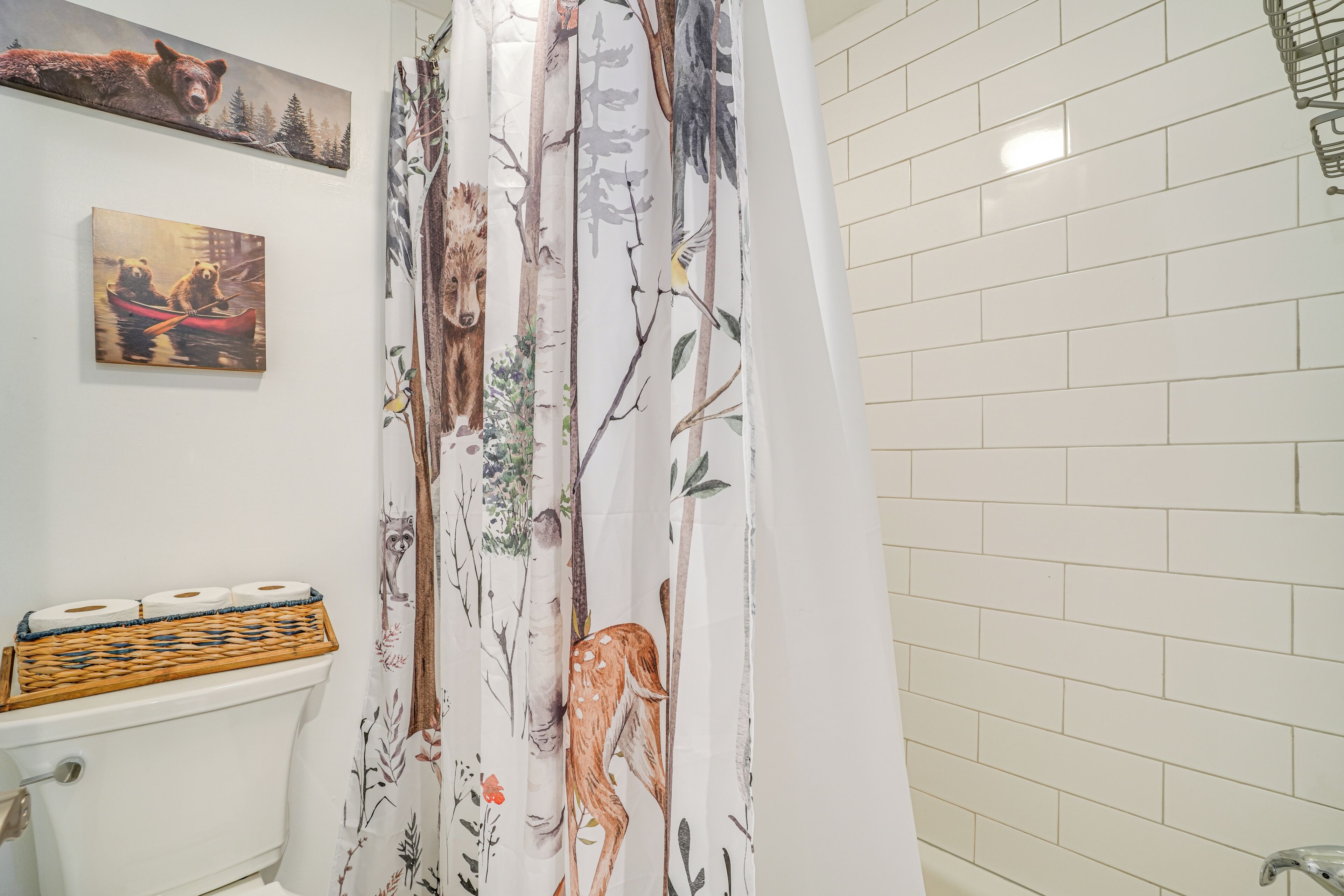 A bathroom with a toilet, a basket of toilet paper, a shower curtain with a forest print, and two wall-mounted paintings.