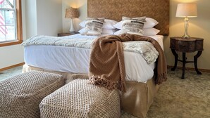 A neatly made bed with a brown headboard, white bedding, and decorative pillows. There are two bedside tables with lamps, a window with a view, and two round ottomans. - < 1 Mi to Bull Shoals Lake: 'the Blue Quill' (Bull Shoals)