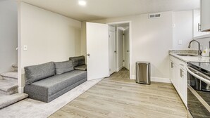 Een modern appartement met een grijze bank, houten vloeren, een keuken met roestvrijstalen apparatuur en een witte deur die naar een andere kamer leidt.