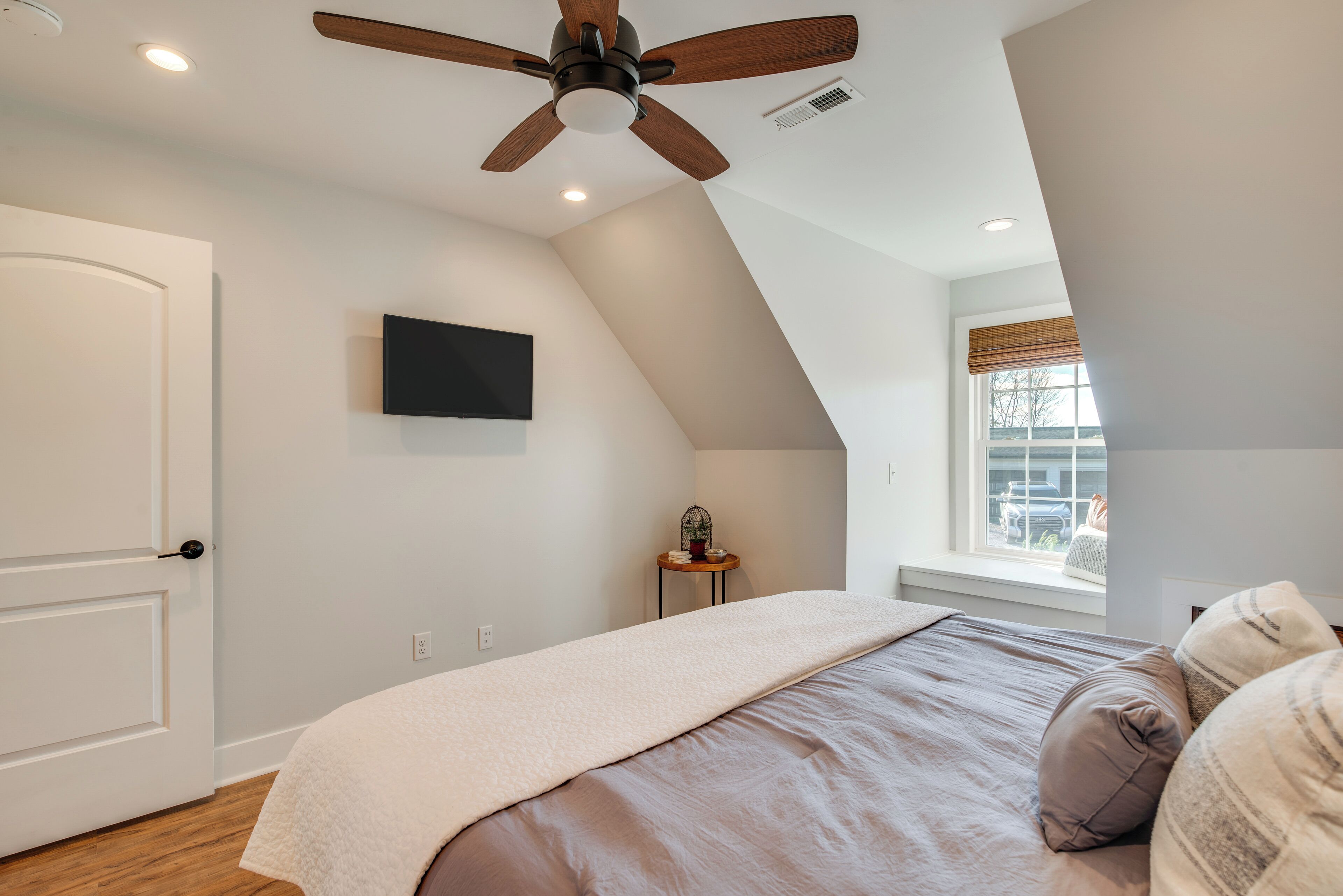 Un dormitorio con techo inclinado, un ventilador de techo, una cama con manta color beige, una mesita con planta y una ventana con persianas.