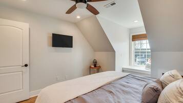 Un dormitorio con techo inclinado, un ventilador de techo, una cama con manta color beige, una mesita con planta y una ventana con persianas.