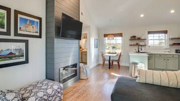 A modern living room with a fireplace, a dining area, and a kitchen with white cabinets and a window.