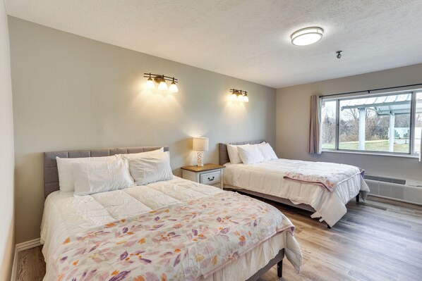 A bedroom with two beds, a nightstand with a lamp, and a large window providing natural light. - Walk to Oakland City University: Modern Apartment! (Oakland City)