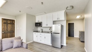 A modern kitchen with white cabinets, a microwave, and a refrigerator. There is a wooden door and a gray sofa in the living area. - < 1 Mi to Oakland City University: Cozy Apartment (Oakland City)