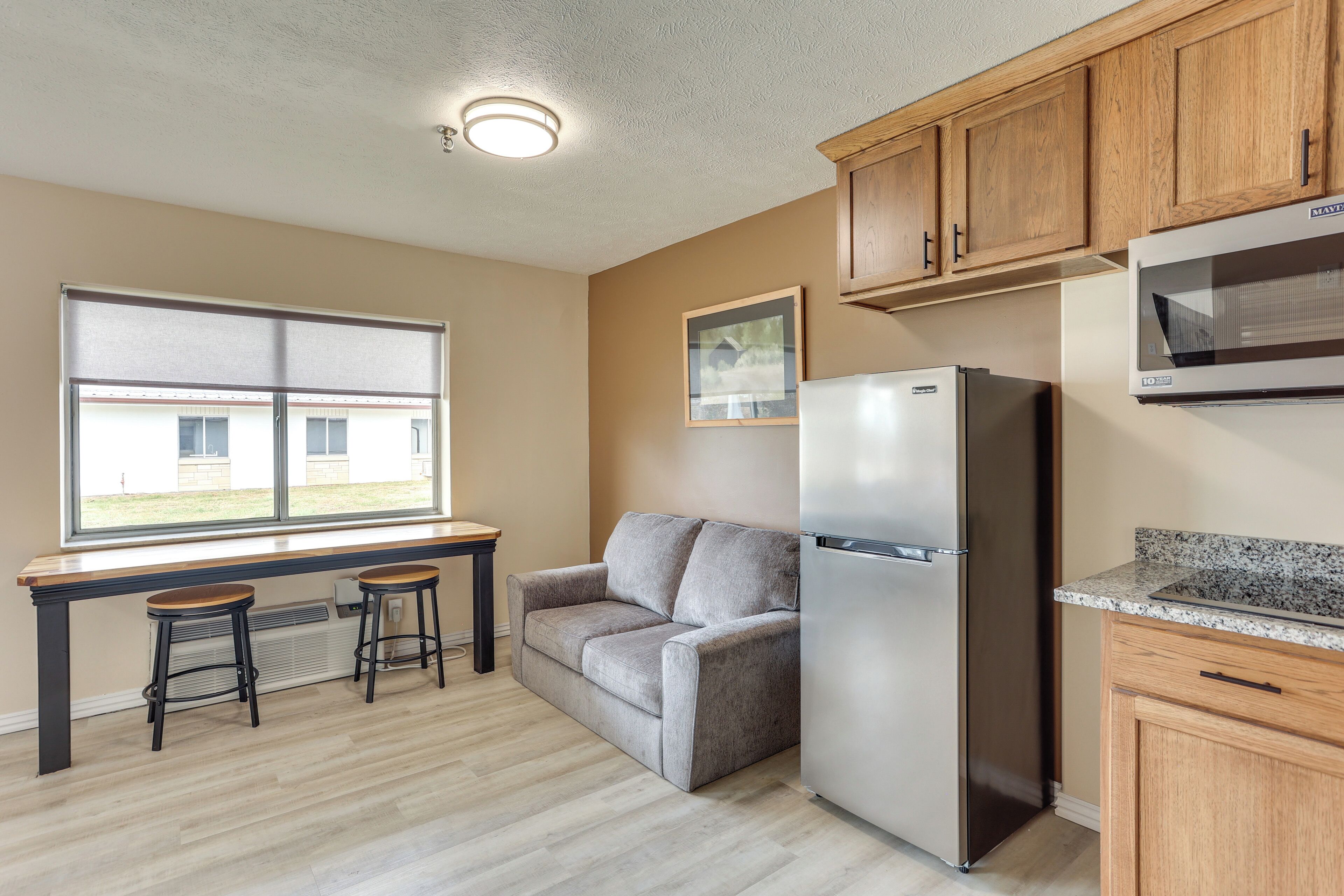 A kitchen with a refrigerator, wooden cabinets, a window with blinds, and a gray sofa with two stools.