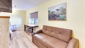 A living room with a brown leather sofa, a dining table with two chairs, a window with a blind, and a painting on the wall. - Quiet Oakland City Studio: Walk Downtown! (Oakland City)