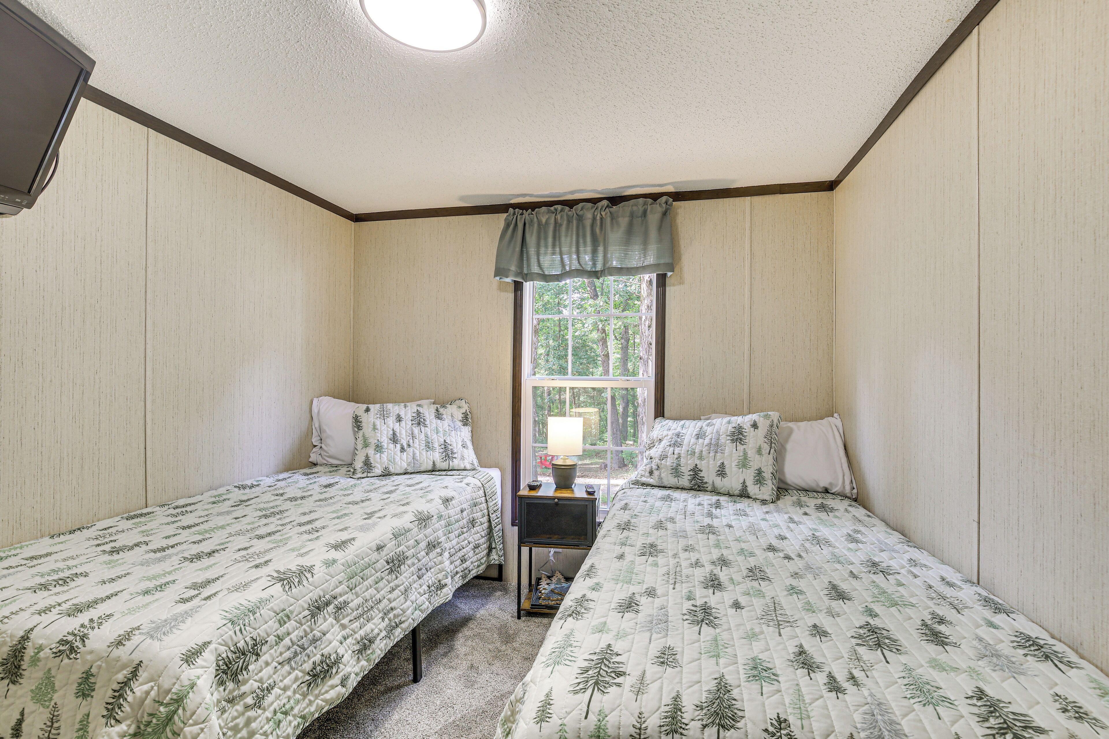 A bedroom with two beds, a nightstand with a lamp, and a window with a view of trees.