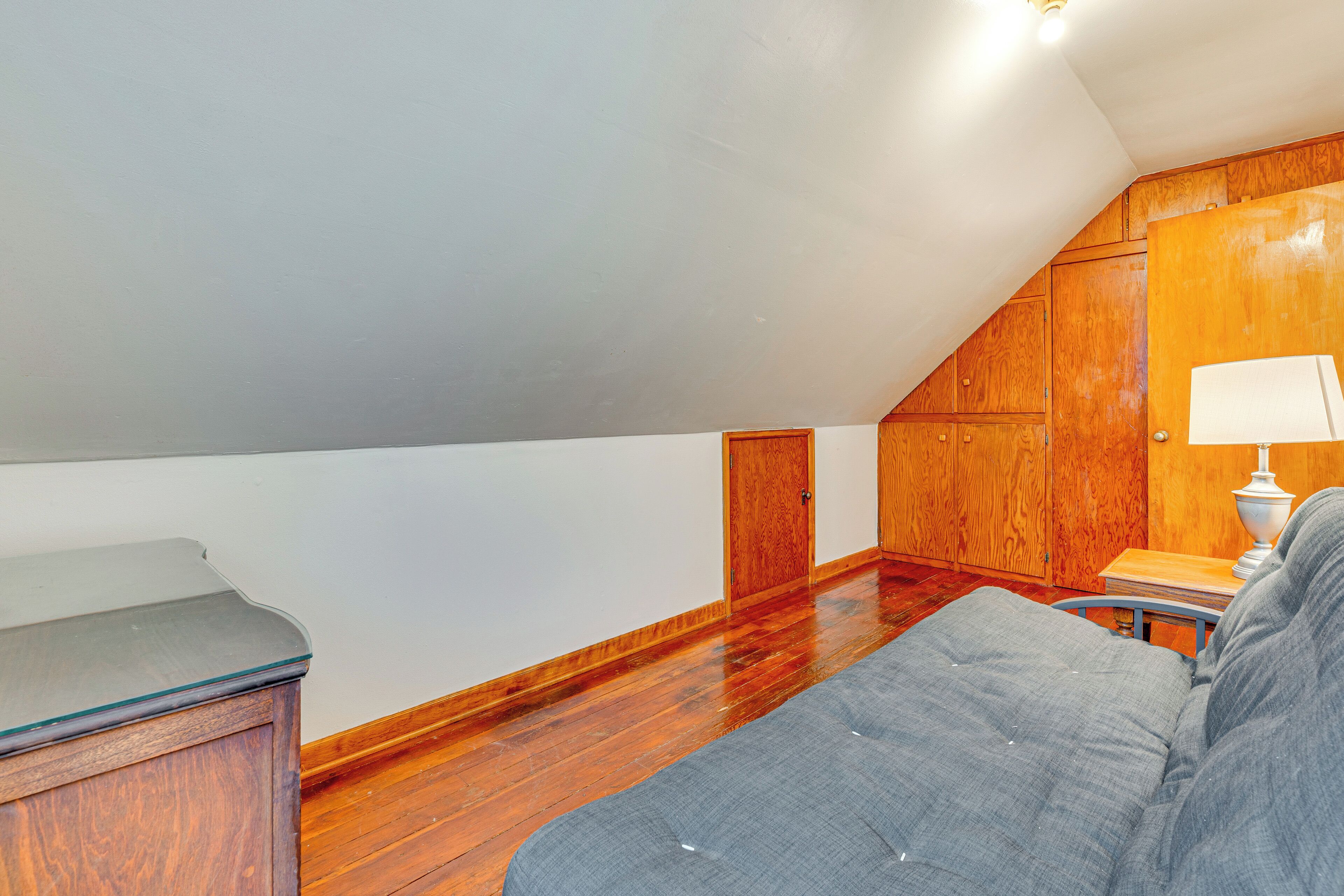 A room with wooden flooring, a grey sofa, a small table with a lamp, and wooden paneling on the walls.