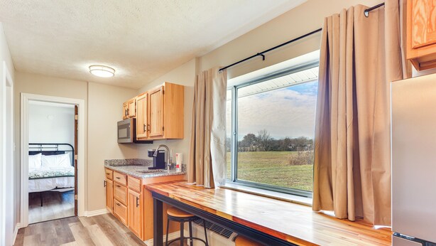 A kitchen with wooden cabinets, a countertop, stools, a microwave, and a large window with curtains. - Golf, Shop, Tour & More: Oakland City Apartment (Oakland City)