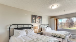A bedroom with two beds, a nightstand with a lamp, a window with a view, and a ceiling light. - Golf, Shop, Tour & More: Oakland City Apartment (Oakland City)