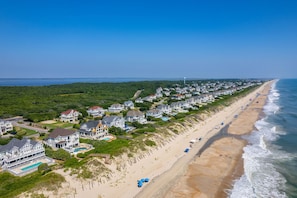 Vue aérienne d’une zone résidentielle côtière, avec des propriétés situées en bord de mer, une longue plage de sable et un ciel bleu dégagé.