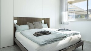 A modern hotel room with a large bed, a wooden headboard, two bedside lamps, and a view of buildings through a window. - Appartamento Guglielmo (Oristano)