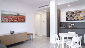 A modern living room with a grey sofa, a dining area with white chairs, a kitchen with a grey countertop, and a wall with a painting of flowers and spices. - Appartamento Guglielmo (Oristano)