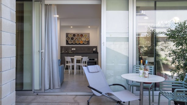 A modern outdoor seating area with a lounge chair, a small round table, and a glass door leading to a kitchen area with a dining table and chairs. - Appartamento Guglielmo (Oristano)