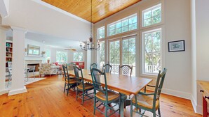 Un vaste espace dédié à la salle à manger, avec une table et des chaises en bois, de grandes fenêtres offrant une vue sur les arbres, et une cheminée en arrière-plan.