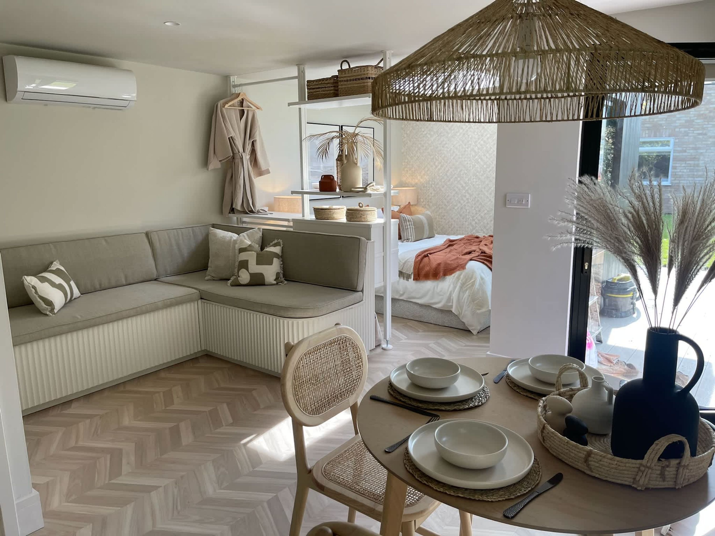 Une pièce moderne et bien éclairée, avec un canapé, une table à manger dressée avec des assiettes et des bols, une lampe en rotin suspendue, et une vue sur une chambre à coucher à travers une porte vitrée.
