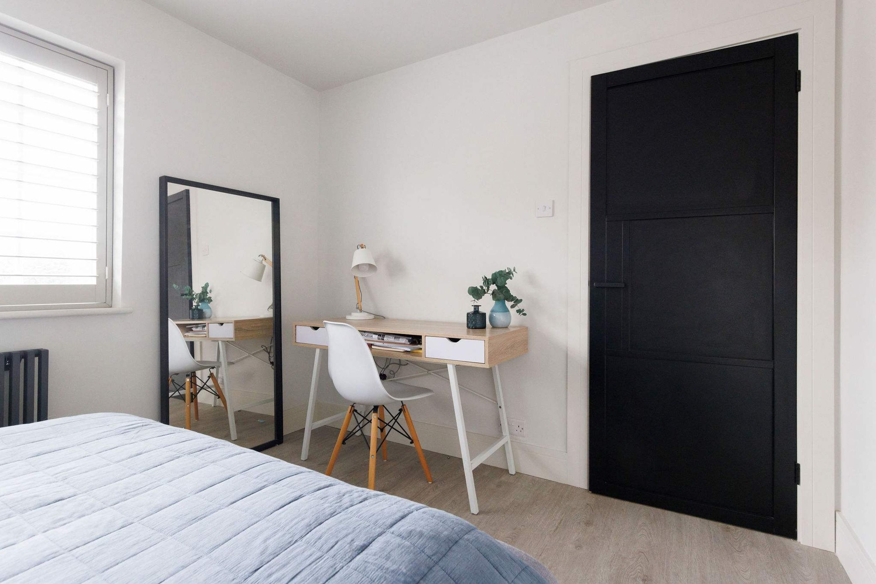 A bedroom with a bed, a desk with a chair, a mirror, and a black door.