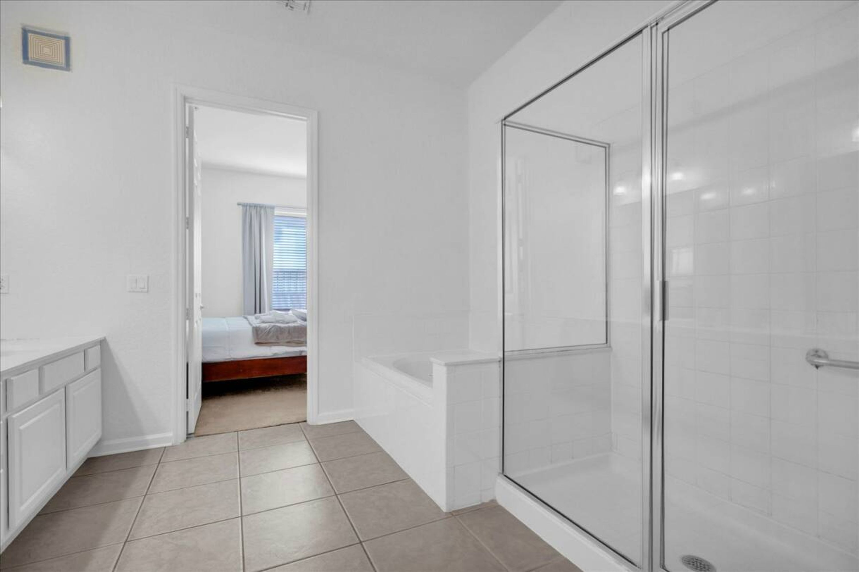 A modern bathroom with a glass shower enclosure, a white bathtub, and a visible door leading to a bedroom with a bed and curtains.