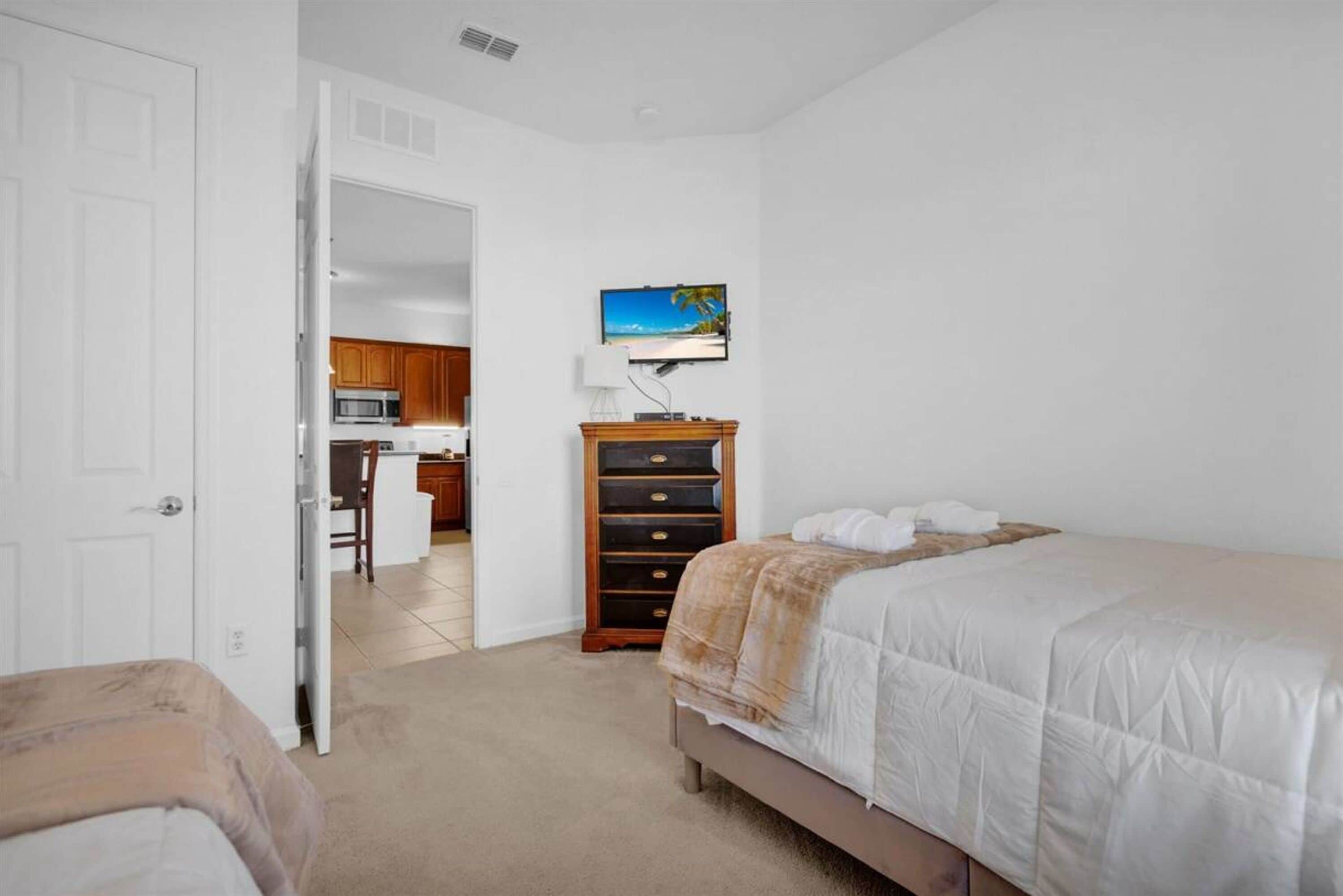 Un dormitorio con cama, cómoda, televisor en la pared y una zona de cocina al fondo.