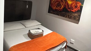 A bedroom with a bed, an orange throw blanket, a painting of roses, and a window with blinds.