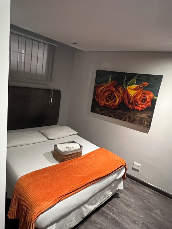 A bedroom with a bed, an orange throw blanket, a painting of roses, and a window with blinds. - Safe and secure Wild olive apartments. 2 minutes walk to the mall and Hospital  (Windhoek)