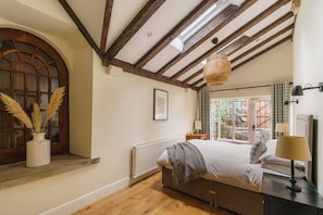 Une chambre avec un plafond en bois, une grande fenêtre avec des rideaux, un lit avec une couverture, une table de chevet avec une lampe et un vase contenant des plumes d’herbe des pampas.