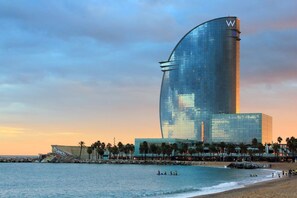 Een modern hotel met een gebogen glazen gevel, nabij een strand met palmbomen en een rotsachtige kustlijn.