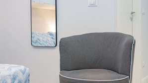 Ein Zimmer mit grauem Polsterstuhl, einem Bett mit gesteppter, blau-weißer Bettdecke und einem wandmontierten Spiegel, der ein Bett und eine Tür reflektiert.