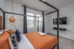 A four-poster bed with a white and orange bedspread, a hanging lamp, a television on the wall, and a window with blinds. - New Construction - Cozy Home & Playroom (Davenport)