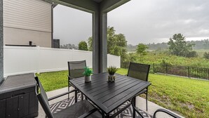 A covered patio with a table and chairs, a white fence, and a view of a grassy area with trees. - New Build - Cozy House & Game Room (Davenport)