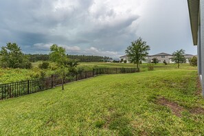 A grassy area with a black metal fence, trees, and a building in the background. - New Construction - Cozy Home & Playroom (Davenport)