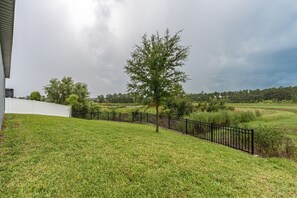 A grassy area with a black metal fence, a white building, and a tree. - New Build - Cozy House & Game Room (Davenport)