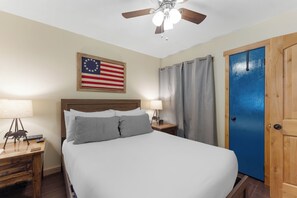 Una habitación de hotel con una cama, una mesita de noche, una lámpara, un ventilador de techo, una puerta azul y una bandera estadounidense en la pared.