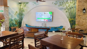 A restaurant with wooden furniture, a large mural of bamboo, and a screen displaying a menu. - Pepitu Dormitory By Sandat (Legian)