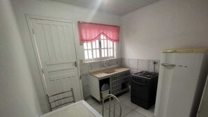 A compact kitchen with a white door, a window with a red checkered valance, a gas stove, a white refrigerator, and a small table with a chair. - Pousada da Pedra do Mar (Matinhos)
