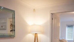 Una habitación con piso de madera, una lámpara de pie, una cama con ropa blanca y un cuadro en la pared.