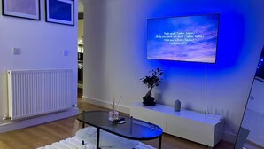 Una sala de estar moderna con un televisor de pantalla plana montado en la pared, una mesa de centro de vidrio, una consola blanca y una planta en maceta.
