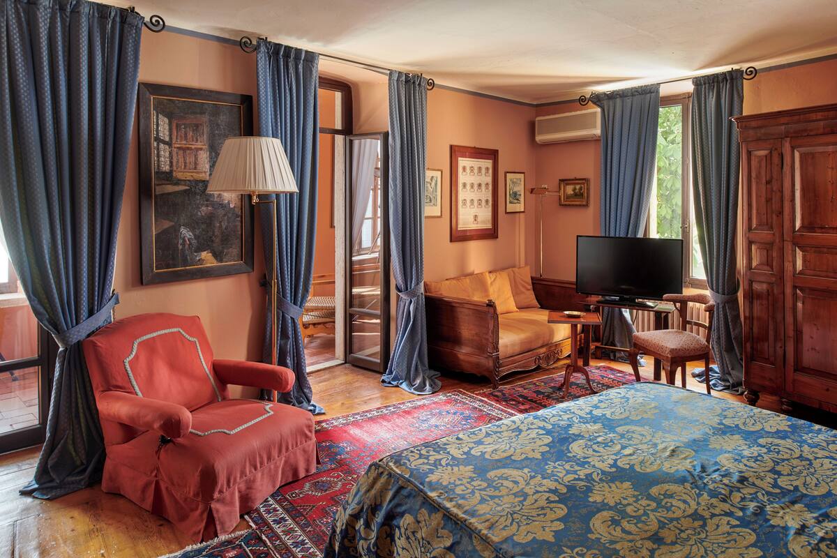 A room with a bed, a red armchair, a sofa, a television, and a large window with curtains.