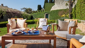 Una mesa de madera servida con alimentos para el desayuno y una botella de champán, rodeada de sillas con cojines, en un jardín con un muro de piedra y follaje al fondo.