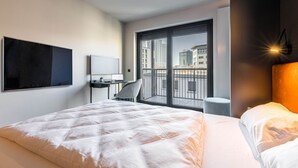 Habitación de hotel moderna con una cama grande, un escritorio con una computadora portátil, una silla y vistas de la ciudad a través de un ventanal.