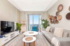 A modern living room with a flat-screen TV, two round tables, a sofa, and a large window offering a city view.