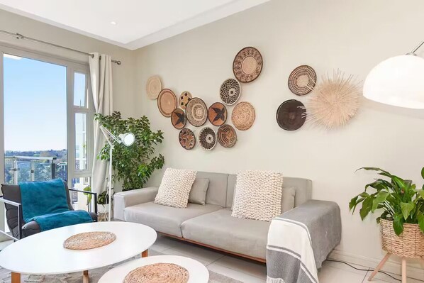 A modern living room with a grey sofa, two white round tables, and a variety of woven wall art.
