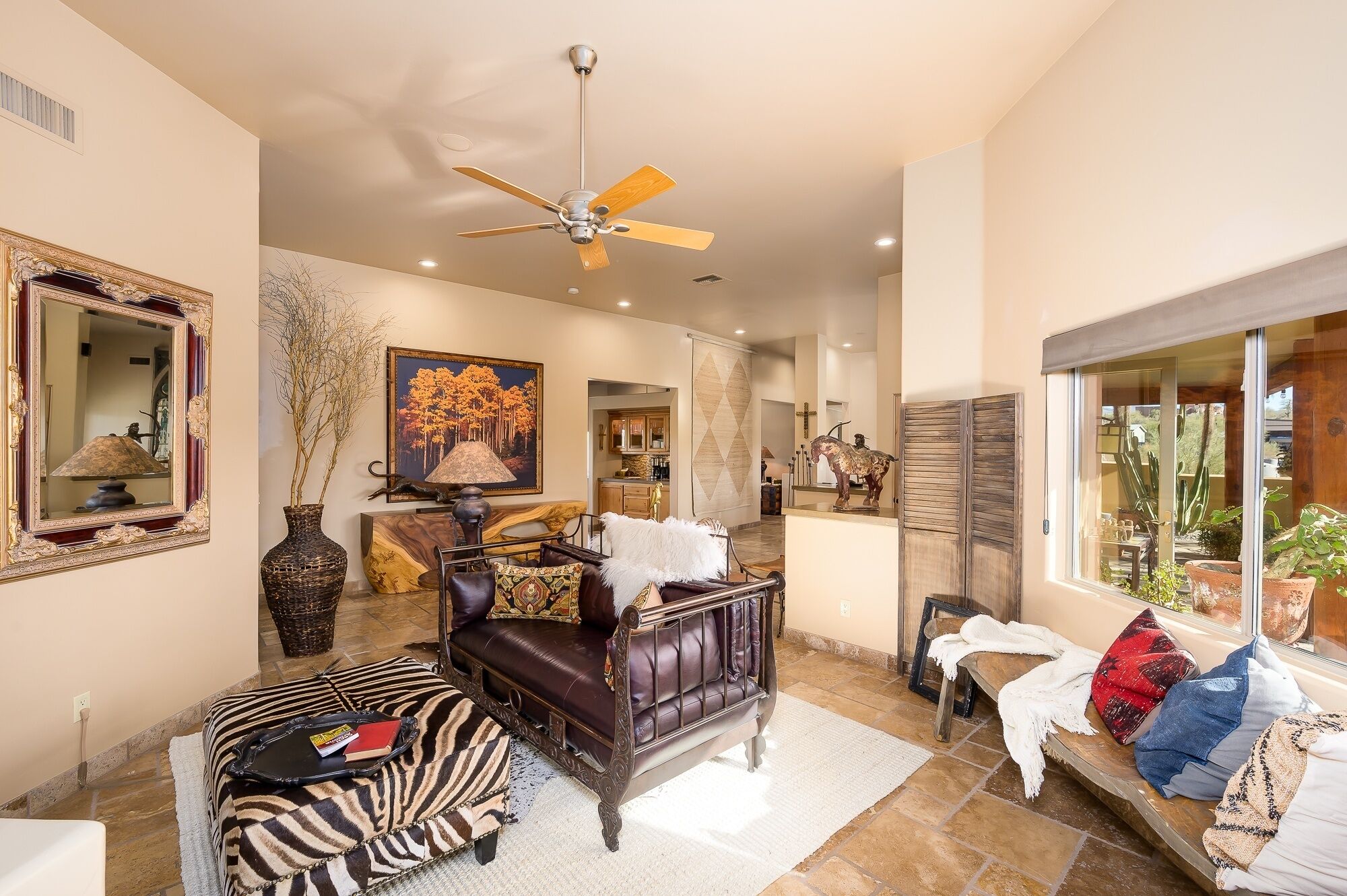 A spacious living room with a ceiling fan, a large mirror, a zebra-patterned ottoman, a leather sofa, and a window with a view of the outdoors.