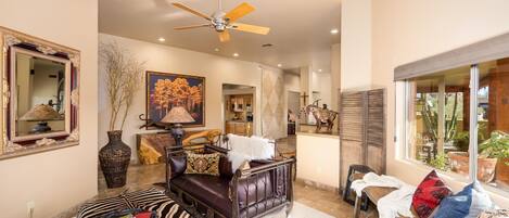 A spacious living room with a ceiling fan, a large mirror, a zebra-patterned ottoman, a leather sofa, and a window with a view of the outdoors.