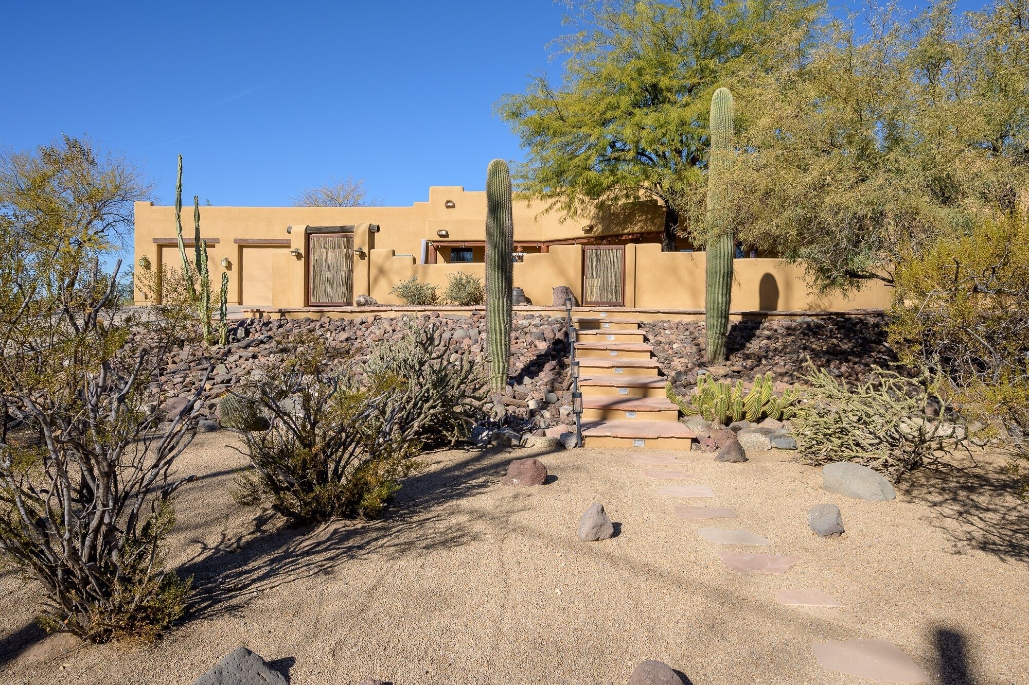 Un bâtiment de style désertique, avec un escalier en pierre menant à une porte, le tout entouré de cactus et d’arbustes.
